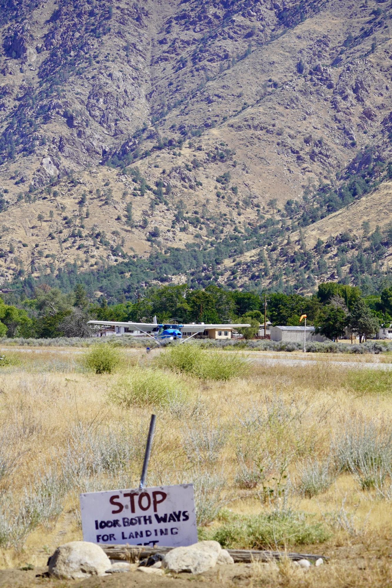 Kern Valley Flyout A hand-painted 'STOP, look both ways for landing' sign in the grass next to the Kern Valley runway, with a parked Cessna and the canyon walls behind it.
